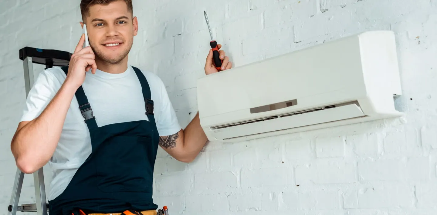 An engineer on the phone with a screwdriver in hand beside an air conditioning unit